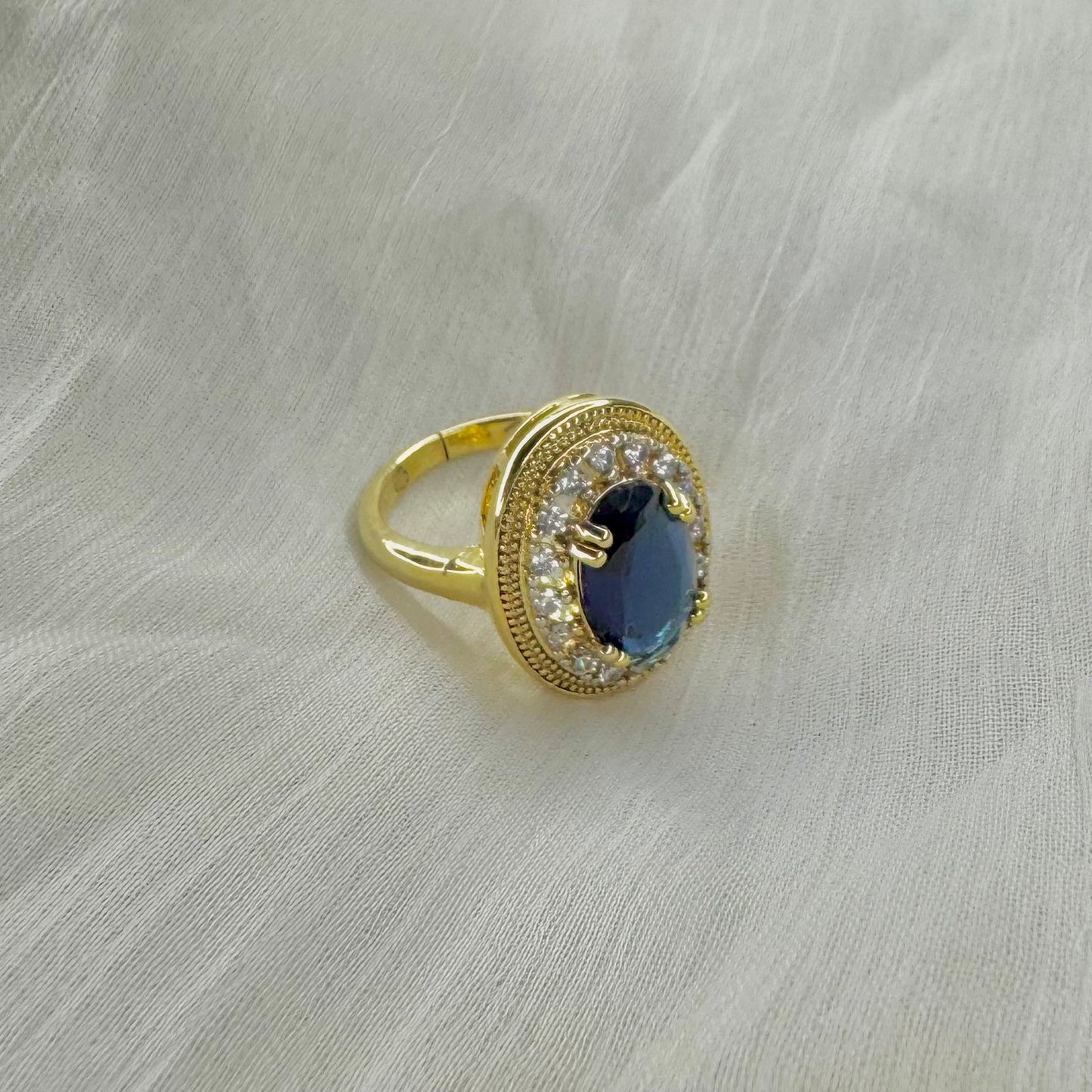 Gold ring with large oval blue sapphire center surrounded by small clear diamonds on cream fabric